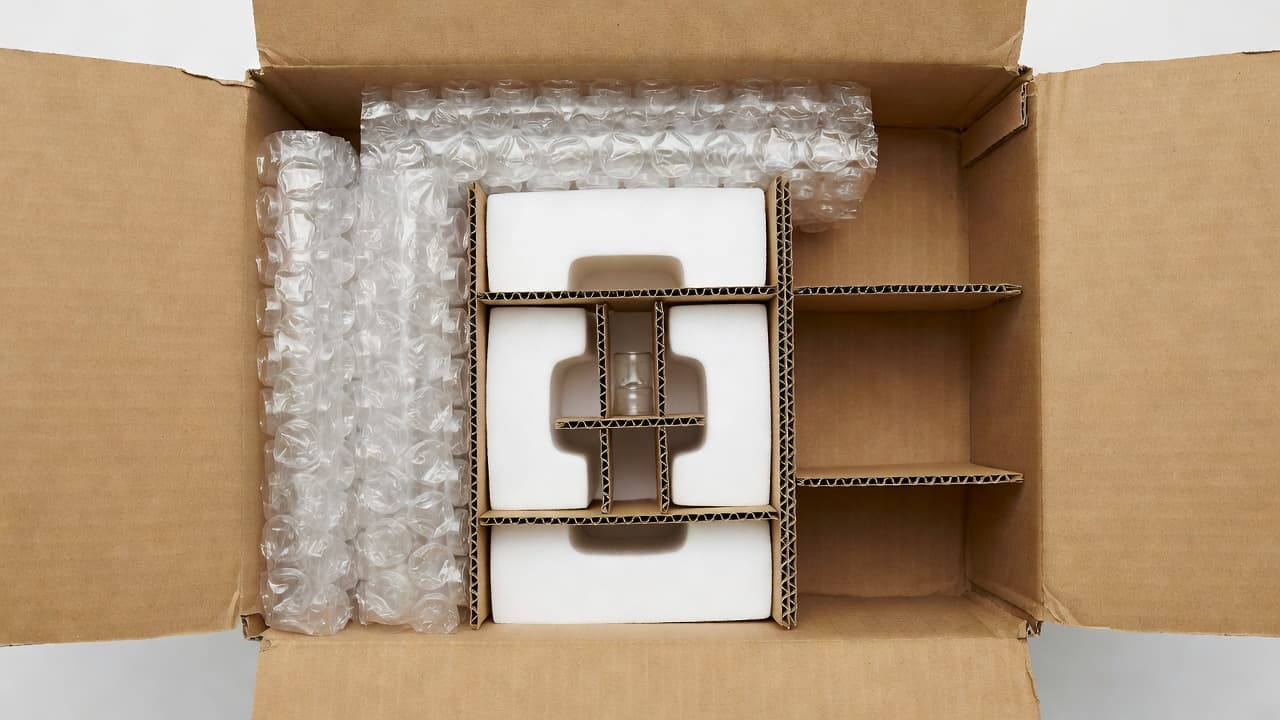 A top-down view shows the interior of a cardboard shipping container featuring custom foam inserts, bubble cushioning, and corrugated paper dividers. These materials safely cradle a small fragile object during transit.